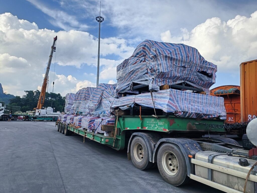 Nhân viên thực hiện chằng buộc và cố định hàng hóa trên mooc lùn bằng xích, tăng đơ và gối chèn trước khi vận chuyển từ Tây Ninh đi Quảng Nam an toàn. chang-buoc-va-co-dinh-hang-tren-mooc-lun-truoc-khi-van-chuyen-di-quang-nam