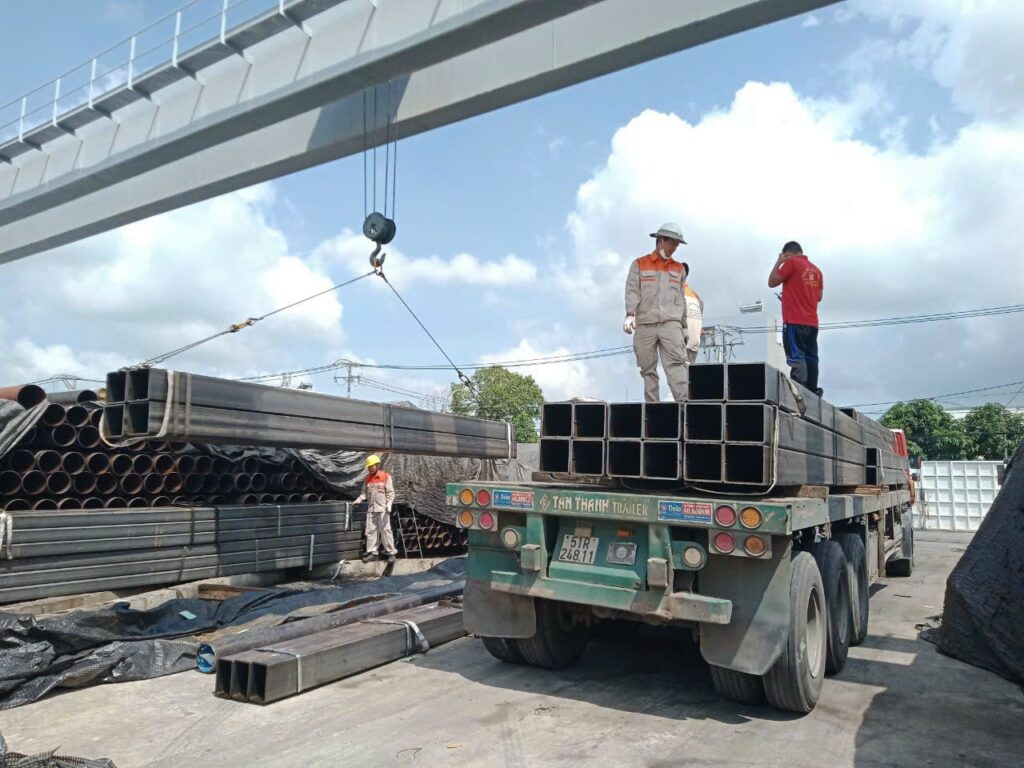 Cẩu các bó thép hộp từ xe đầu kéo mooc sàn xuống kho, dùng thiết bị nâng hạ để di chuyển hàng hóa và sắp xếp đúng vị trí. cau-ha-cac-bo-thep-hop-tu-xe-mooc-san-xuong-kho-hang